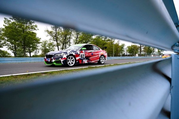 Race car with number 62 speeding along a track, framed by blue safety barriers and green trees in the background.